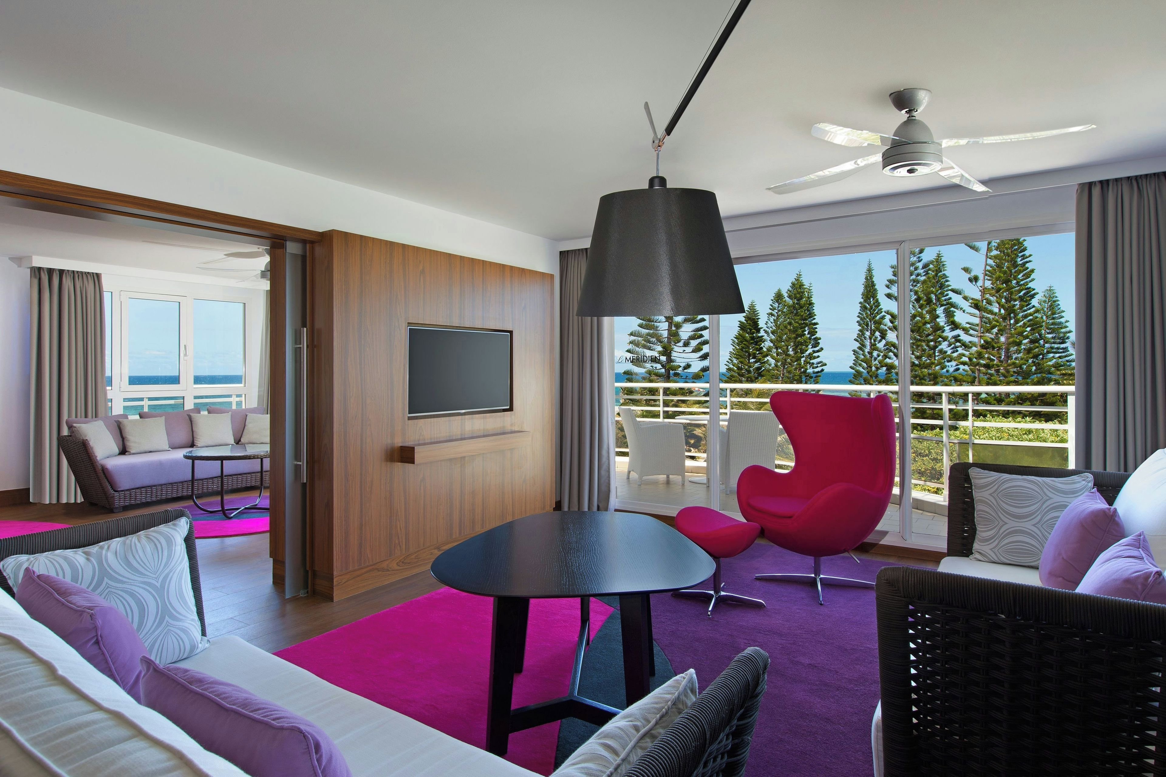 A modern hotel room with a large window, a ceiling fan, a flat-screen TV, and a balcony with outdoor furniture.