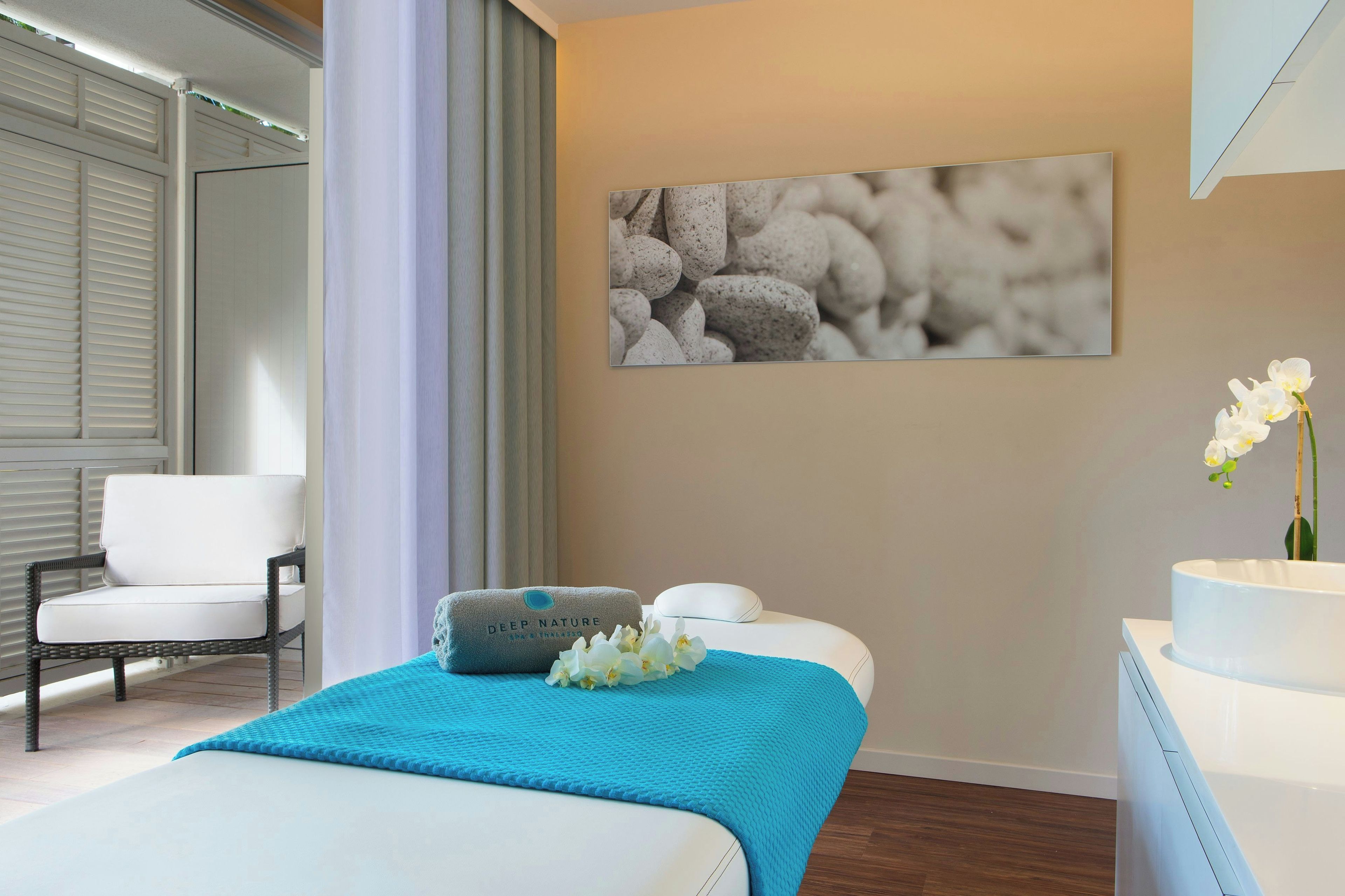 A hotel room with a bed, a chair, a massage table, a wall-mounted picture, and a vase with flowers.