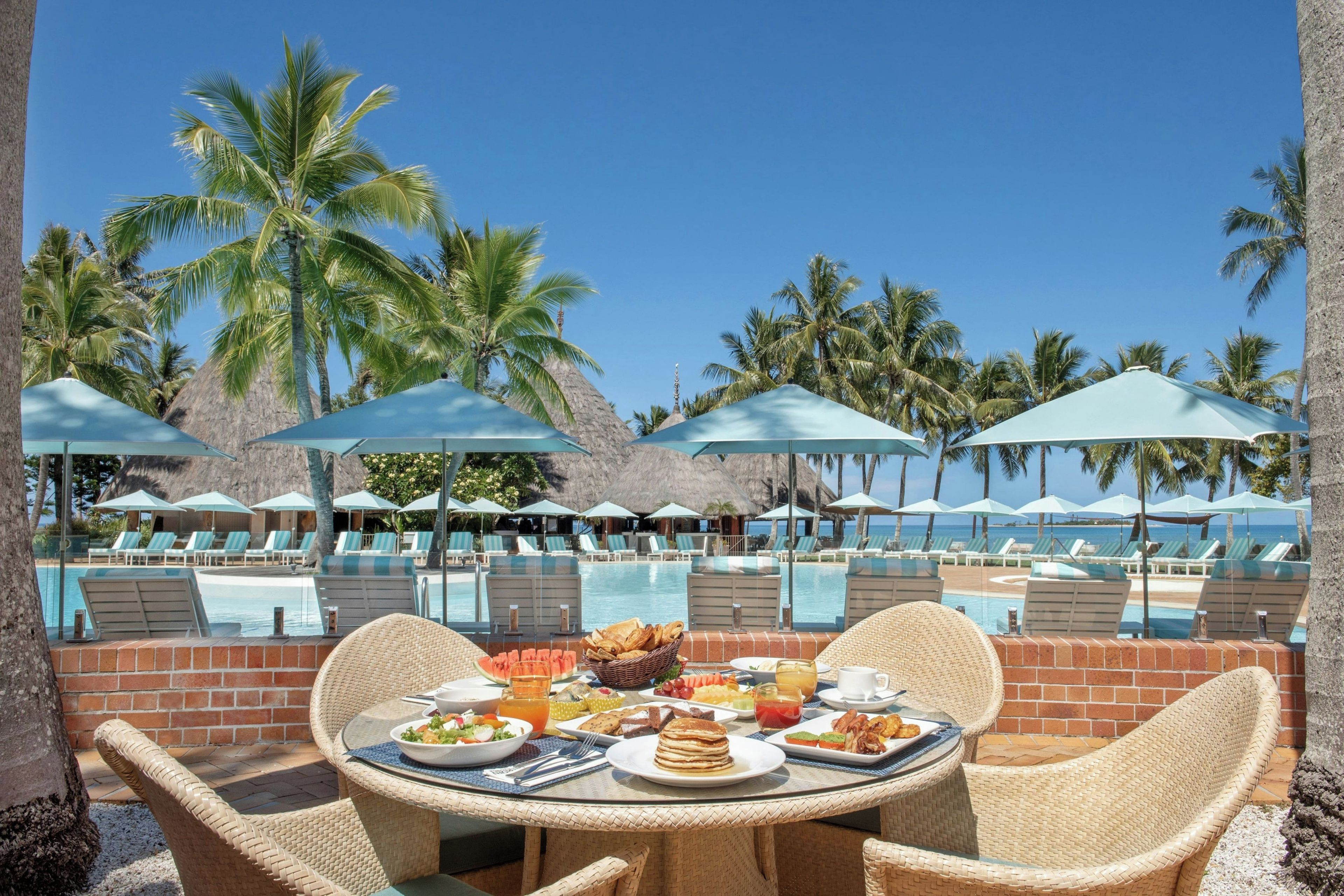 Un área de comedor junto a la piscina con una mesa dispuesta para el desayuno, con vistas a una piscina con sillas de descanso y sombrillas.