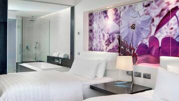 A modern hotel room with a large bed, a bedside table, a mirror, a bathroom with a glass shower enclosure, and a decorative wall with floral patterns.