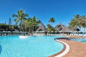 A swimming pool with lounge chairs and umbrellas, surrounded by palm trees and thatched-roof structures.