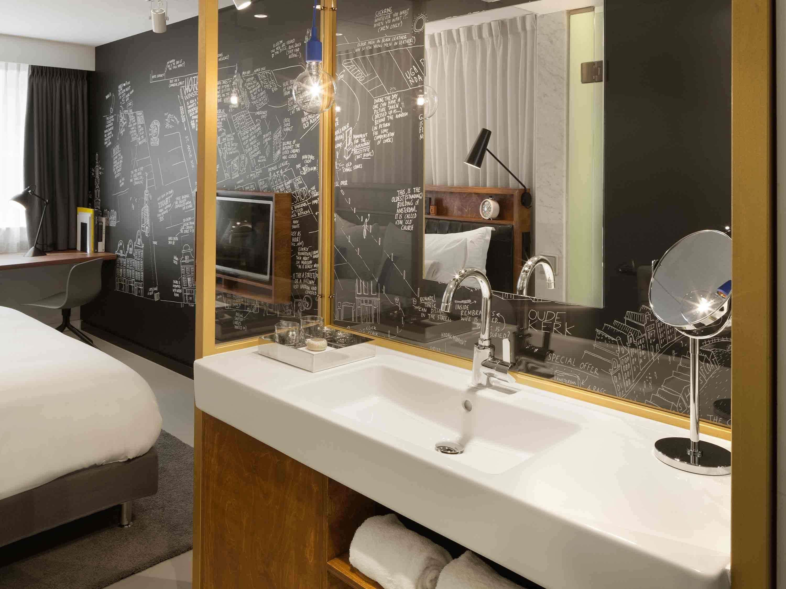 Ein modernes Hotelzimmer mit einem großen Spiegel, einem weißen Waschbecken und einem Bett mit Holz Kopfteil.