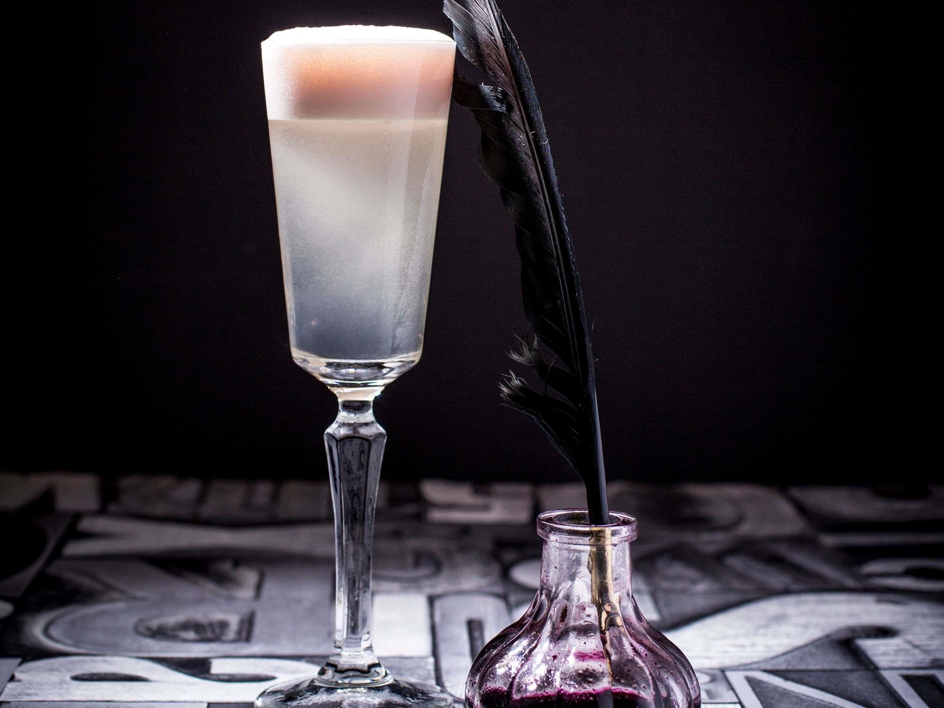 A tall glass with a layered drink, a feather quill, and a small bottle on a patterned surface.