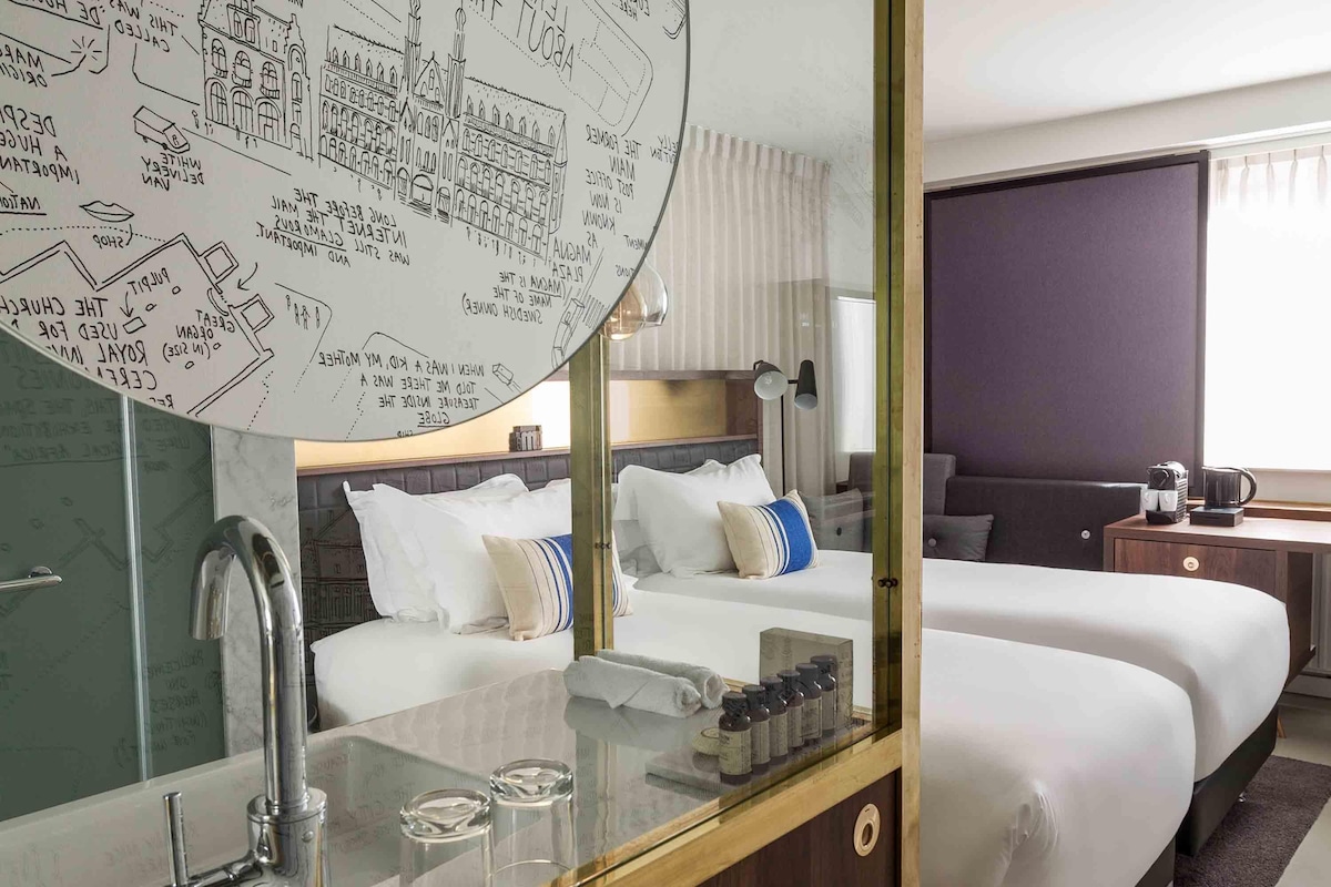 A hotel room with a large mirror, a bed, a bedside table with a lamp, and a window.