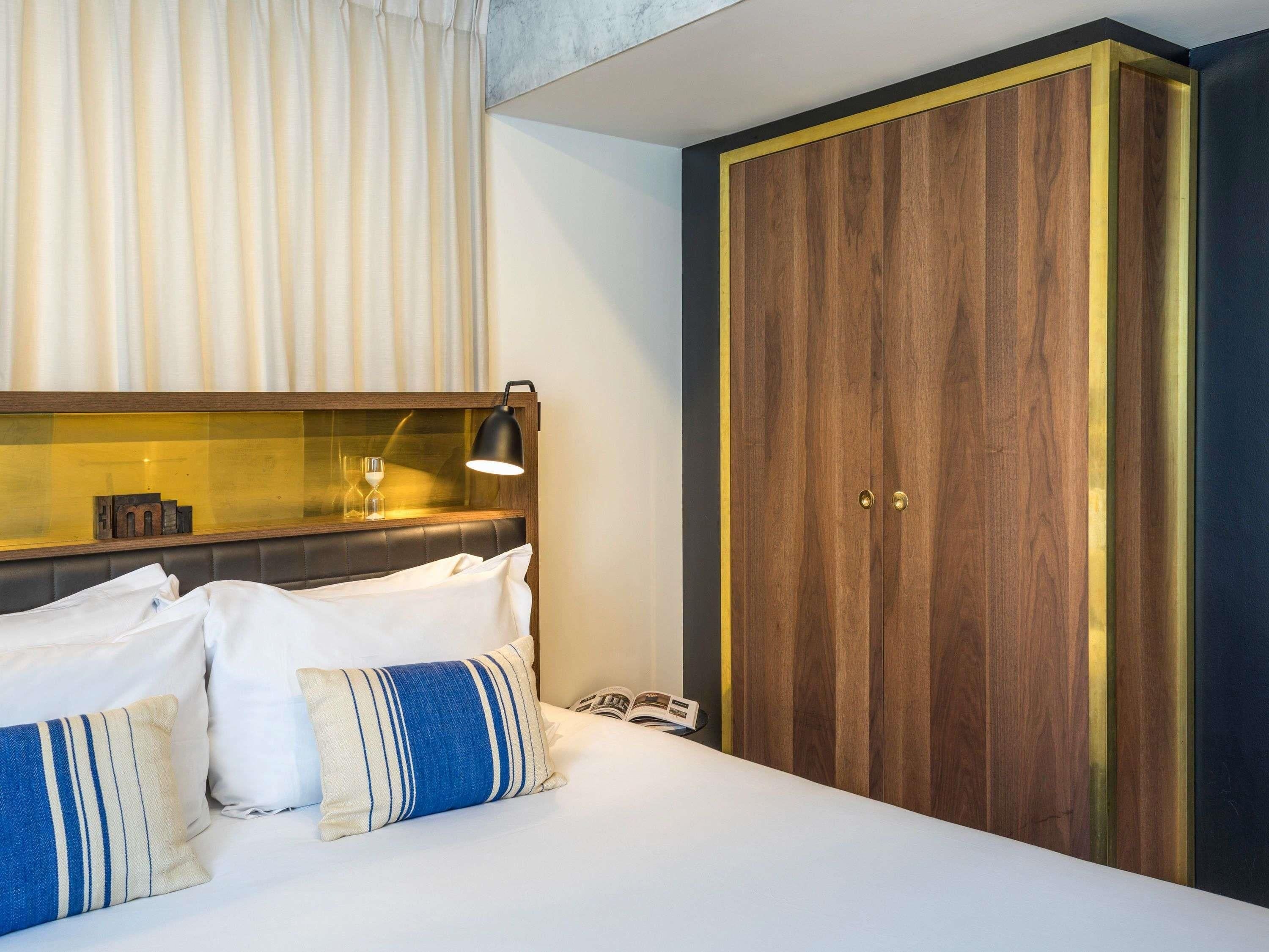 A hotel room with a bed, a bedside lamp, a wooden wardrobe, and a mirror above the bed.