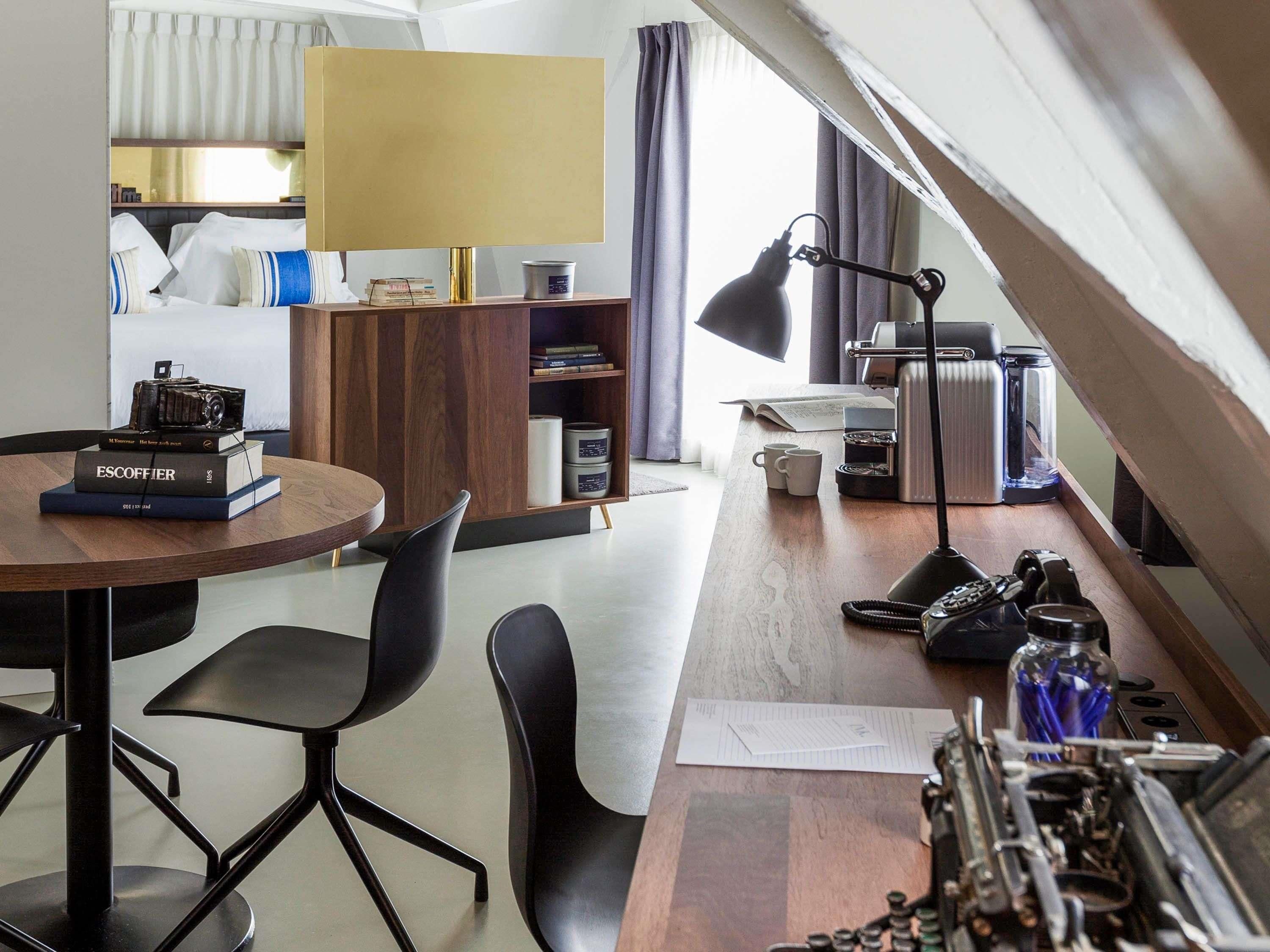 Une chambre moderne avec un bureau en bois, des chaises noires, une bibliothèque, une lampe, une machine à café et un radiateur.