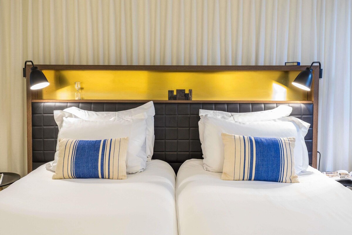 A hotel room with a bed, two bedside lamps, a clock, and a nightstand.