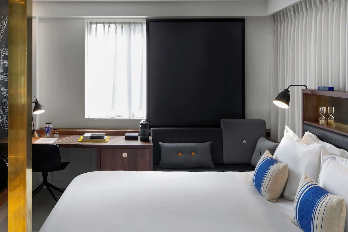 Ein Hotelzimmer mit Bett, Schreibtisch, Stuhl, einem Fenster mit Vorhängen und einem Nachttisch mit Gläsern.