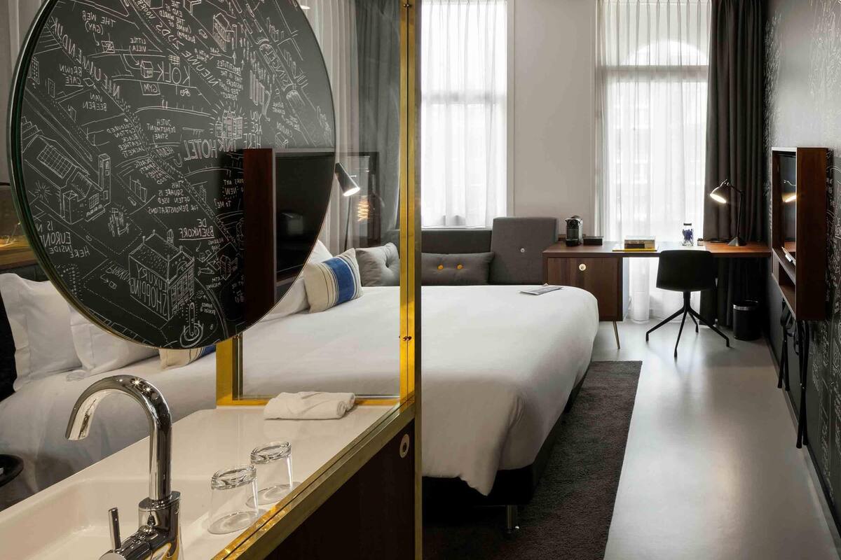 Ein Hotelzimmer mit großem Spiegel, einem Bett, einem Schreibtisch, einem Stuhl und einem Fenster mit Vorhängen.