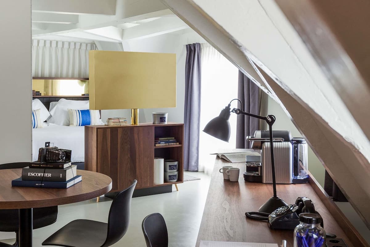 Ein modernes Hotelzimmer mit Schreibtisch aus Holz, Bücherregal, Lampe, Heizkörper und einem Bett mit Vorhängen.