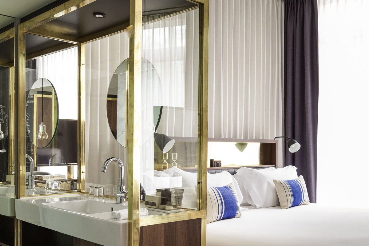 A modern hotel room with a large bathroom vanity, a double bed, and a large mirror.