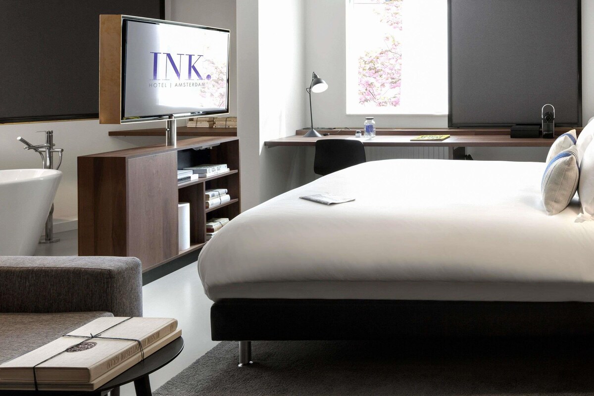 Ein modernes Hotelzimmer mit einem großen Bett, einem Fernseher auf einem Holzständer, einem Schreibtisch mit einer Lampe und einem Fenster mit Blick auf rosa Blumen.