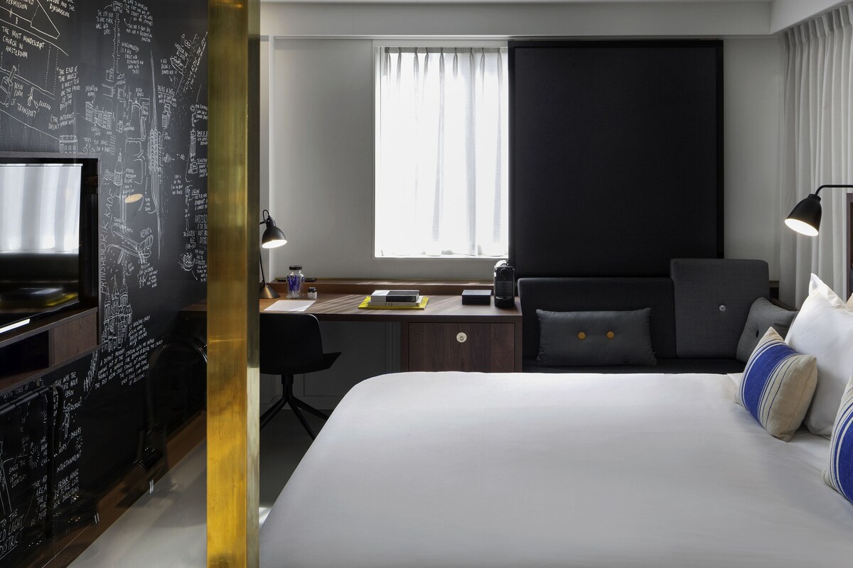 A modern hotel room with a large bed, a desk with a computer, a chair, a TV, and a window with curtains.