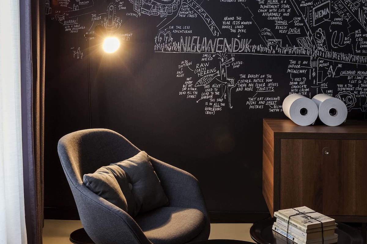 Ein modernes Zimmer mit einer schwarzen Wand, auf der eine Karte und Text im Kreide-Stil dargestellt sind, ein grauer Sessel, ein kleiner runder Tisch mit einem Stapel Bücher und ein Holzschrank mit zwei Toilettenpapierrollen.
