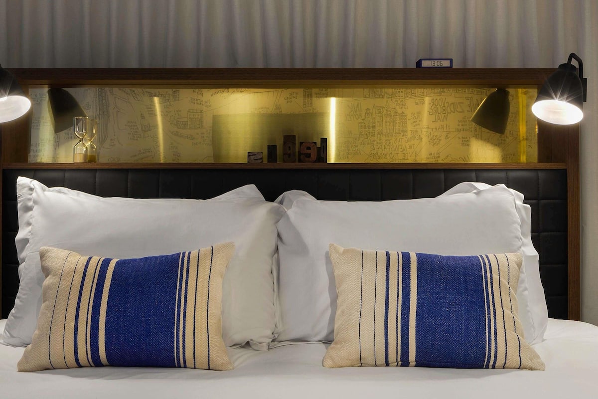 Ein Hotelzimmer mit einem Bett, zwei Dekokissen, einem Kopfteil mit einer Uhr und einer Wand mit einer Weltkarte.