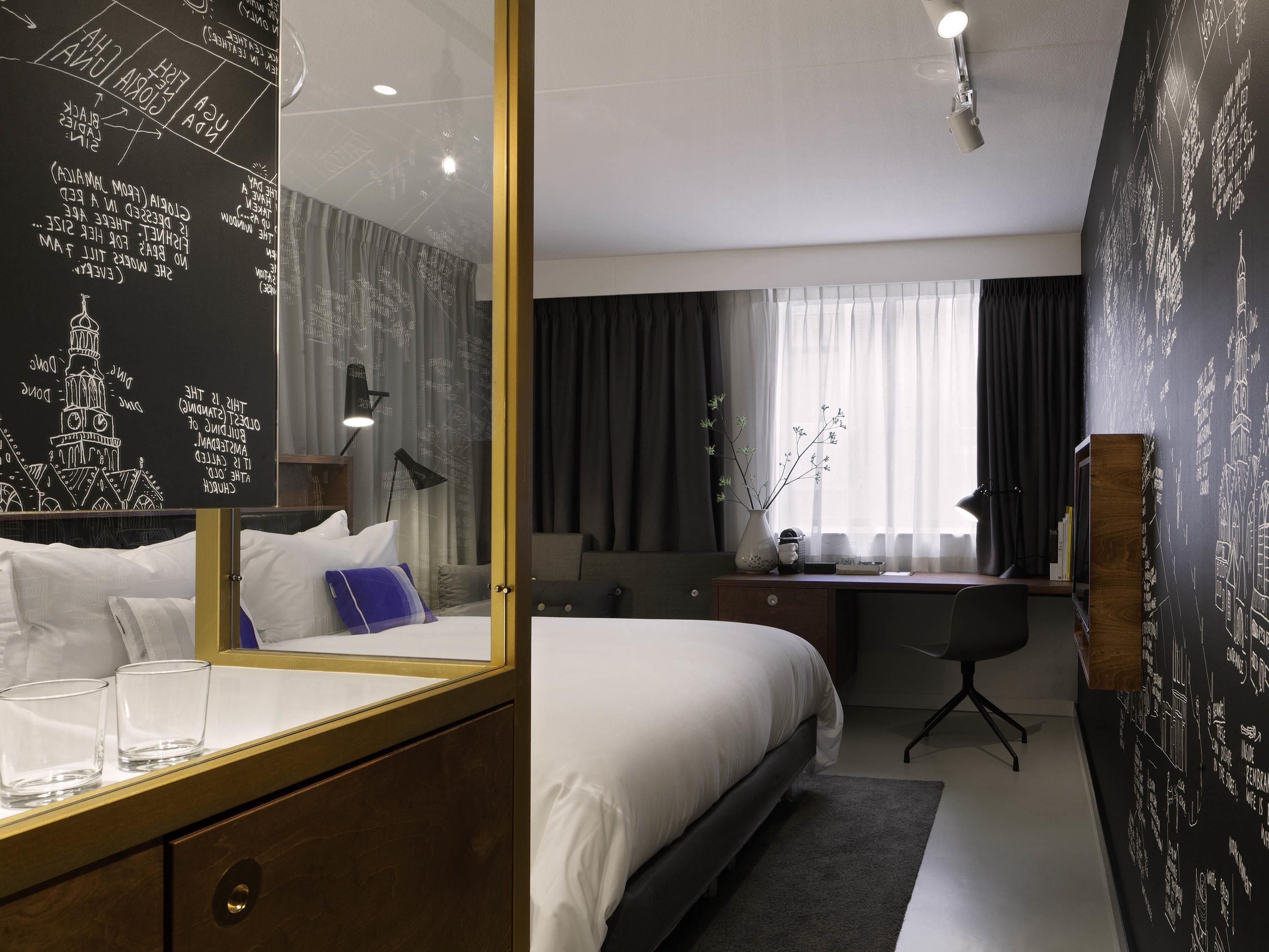 Una habitación de hotel con una cama grande, un escritorio con una silla, una lámpara de escritorio, un jarrón con flores y un espejo amplio.