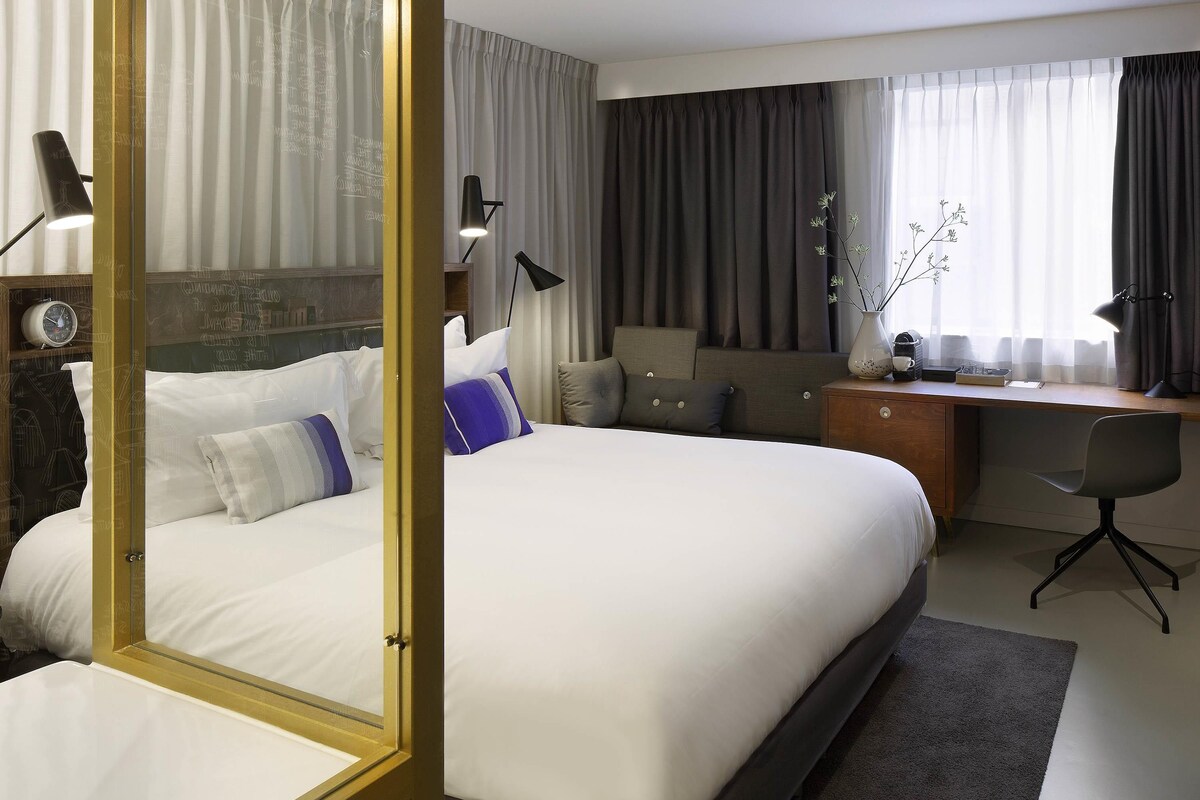 A hotel room with a large bed, a desk, a chair, and a nightstand.