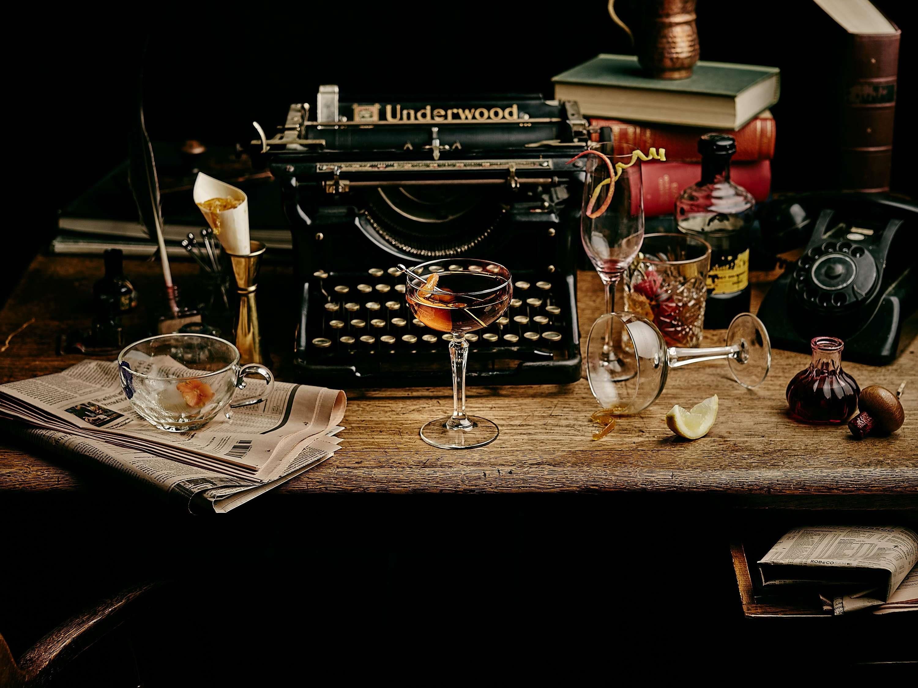 A vintage Underwood typewriter, a glass with a drink, a bottle, and a book on a wooden table.