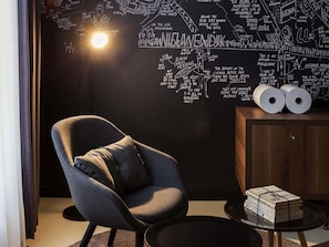 Una habitación con pared negra que presenta un mapa y texto al estilo pizarra, un sillón gris, un armario de madera con papel higiénico y una pequeña mesa redonda.