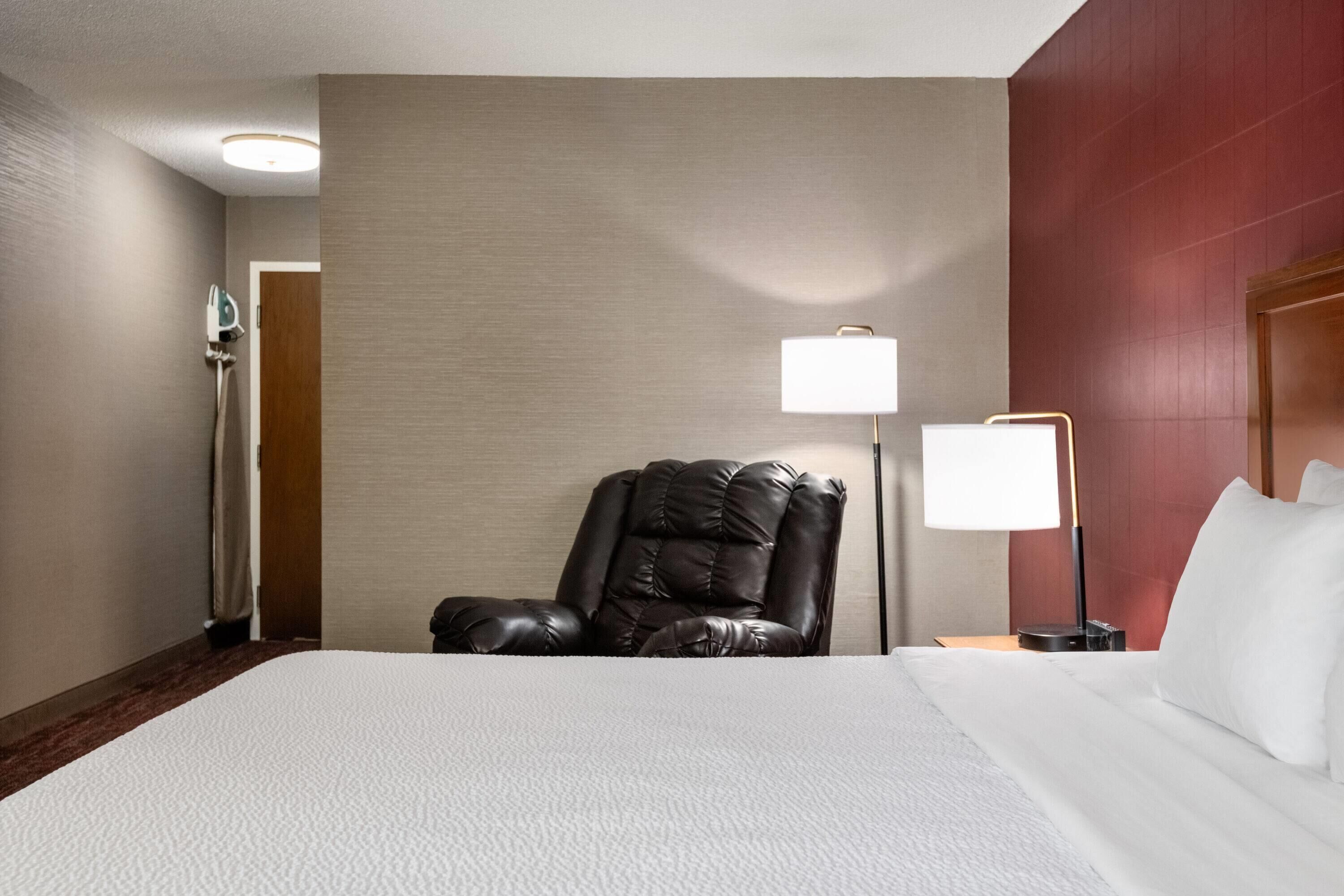 A hotel room with a large bed, a leather chair, a nightstand with a lamp, and a wall-mounted heater.