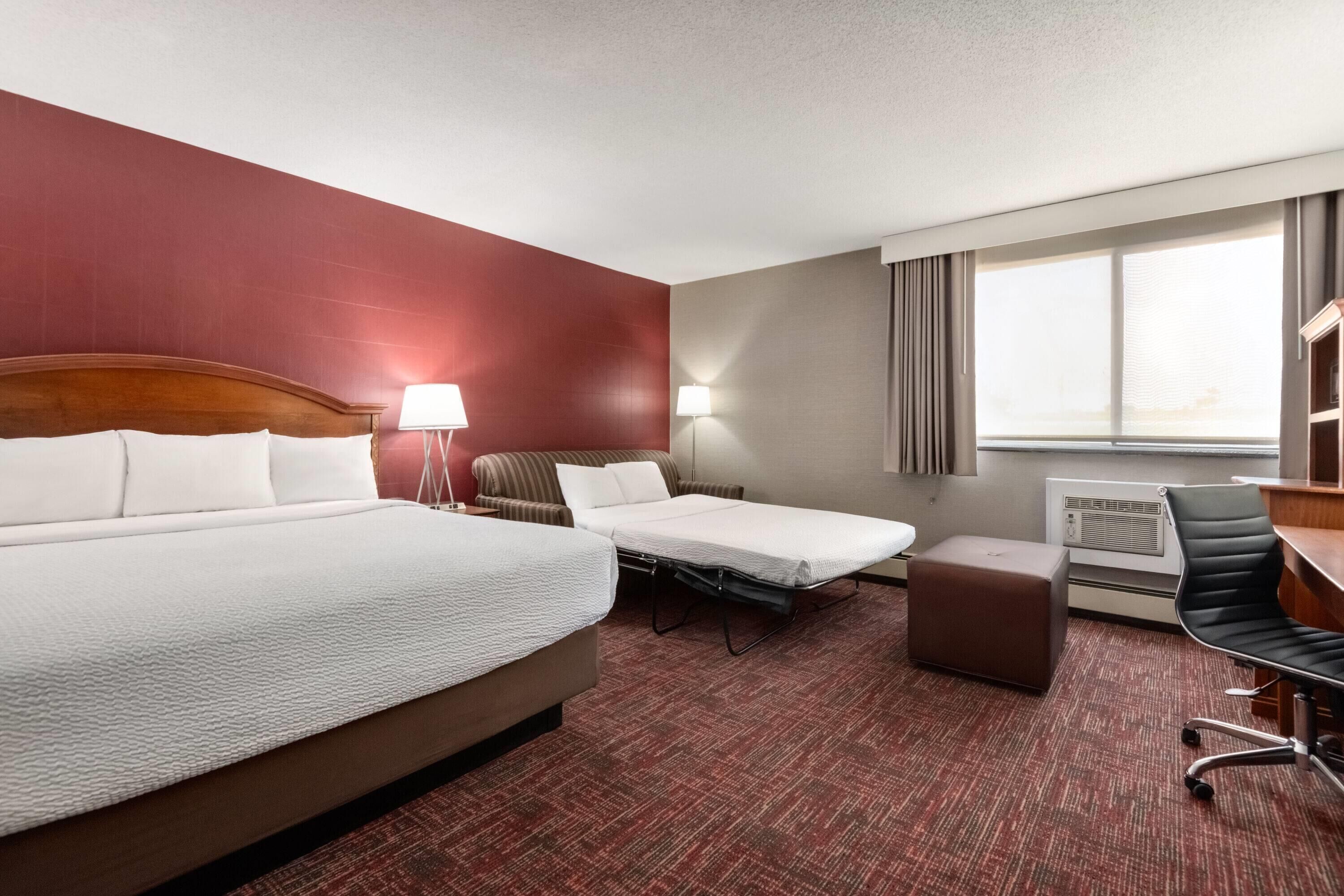 A hotel room with a large bed, a smaller bed, a desk, a chair, a lamp, and a window with curtains.