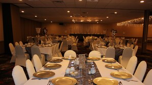 A banquet hall with multiple round tables set with gold-rimmed plates, white tablecloths, and white chairs. - Ramada by Wyndham Grand Forks (Grand Forks)