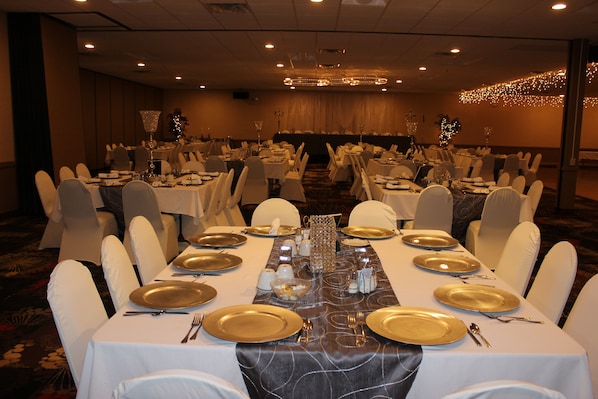 A banquet hall with multiple round tables set with gold-rimmed plates, white tablecloths, and white chairs. - Ramada by Wyndham Grand Forks (Grand Forks)