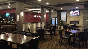A restaurant with a bar area, multiple tables and chairs, and a television screen mounted on the wall. - Ramada by Wyndham Grand Forks (Grand Forks)