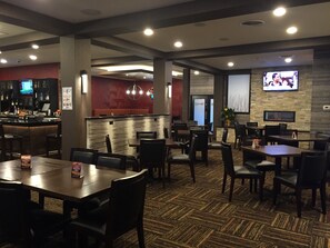 A restaurant with a bar area, multiple tables and chairs, and a television screen mounted on the wall. - Ramada by Wyndham Grand Forks (Grand Forks)