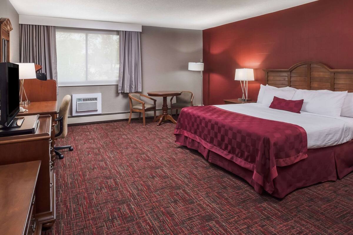 A hotel room with a large bed, a desk with a computer, a chair, a small table, and a window with curtains.