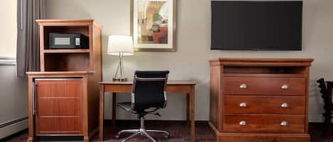 A hotel room with a desk, a chair, a microwave, a cabinet, a TV, and a framed picture on the wall.
