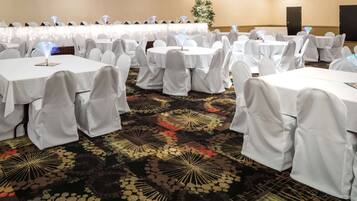 Un salón de banquetes con mesas redondas cubiertas con manteles blancos y sillas, una alfombra estampada y una planta decorativa al fondo.