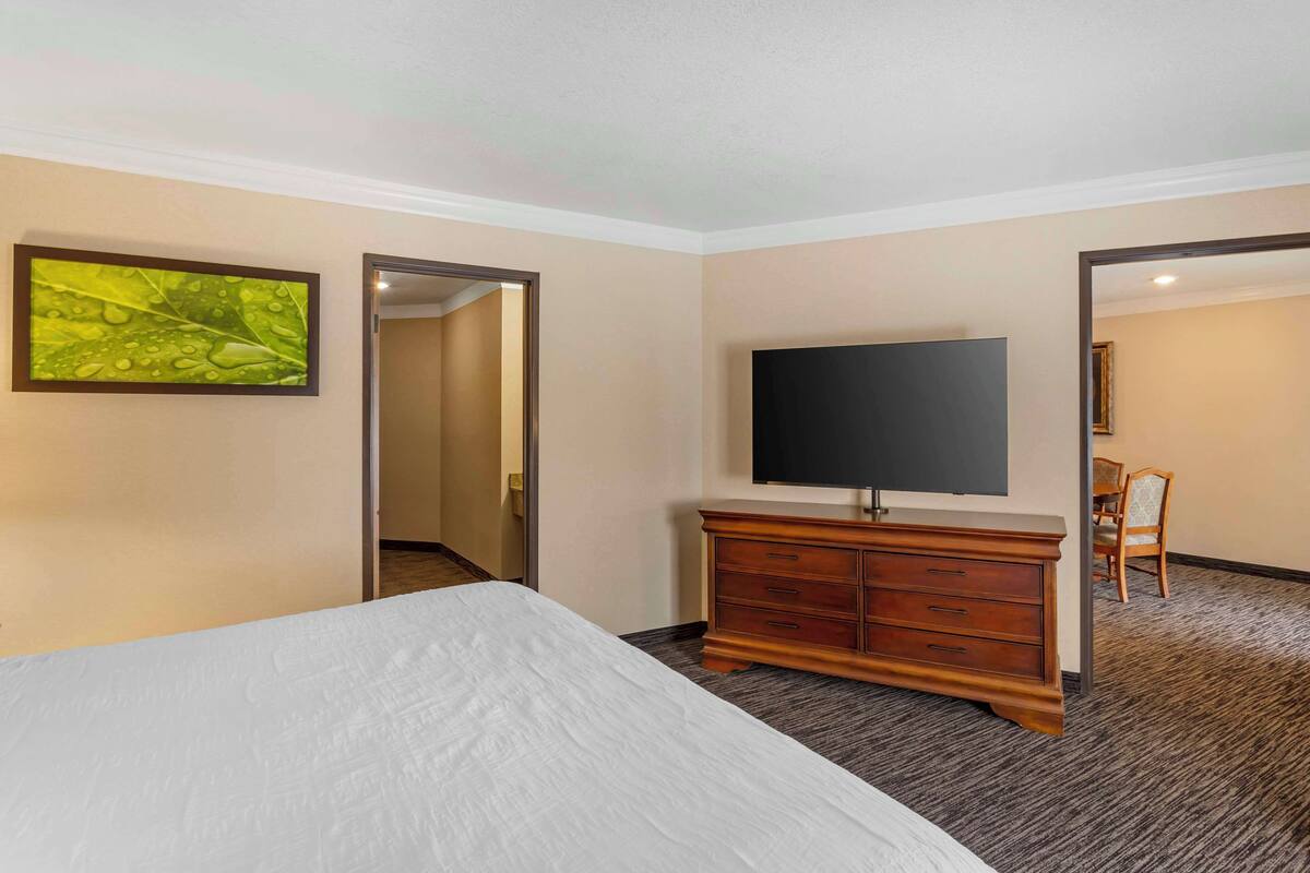 A hotel room with a bed, a flat-screen TV mounted on the wall, a wooden dresser, and a door leading to another room.