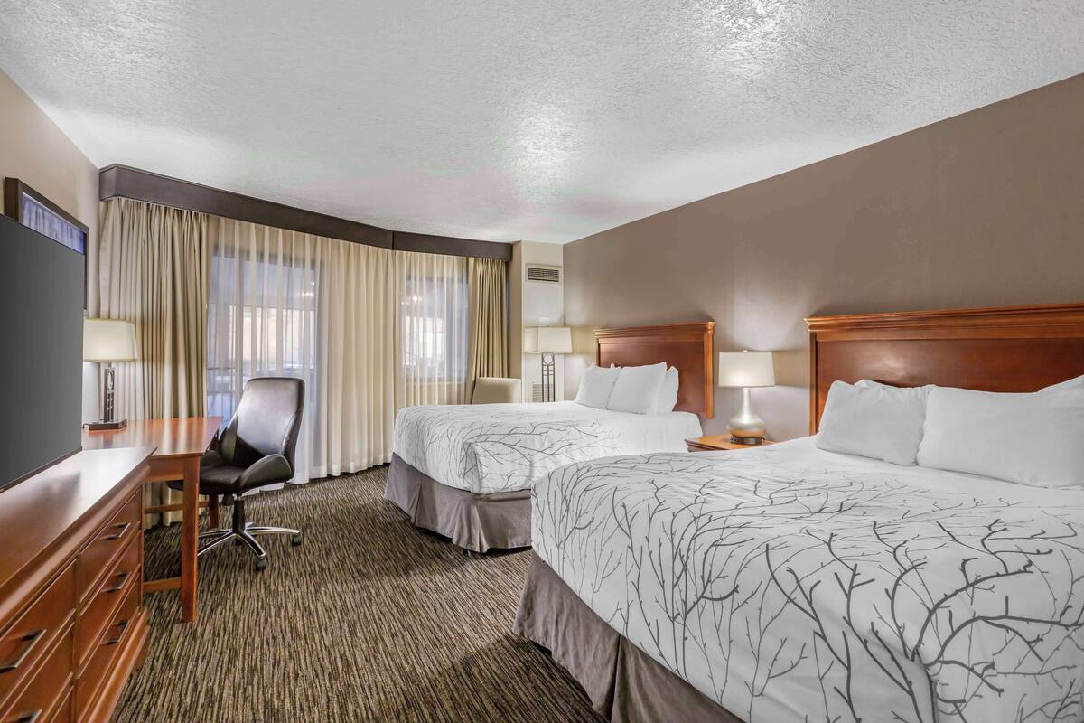 A hotel room with two beds, a desk, a chair, and a large window with curtains.