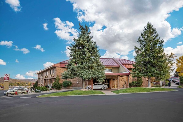 A hotel with a brick facade, a red roof, and two large evergreen trees in front.