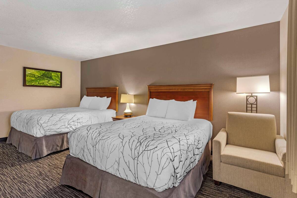 A hotel room with two beds, a chair, a lamp, and a painting on the wall.