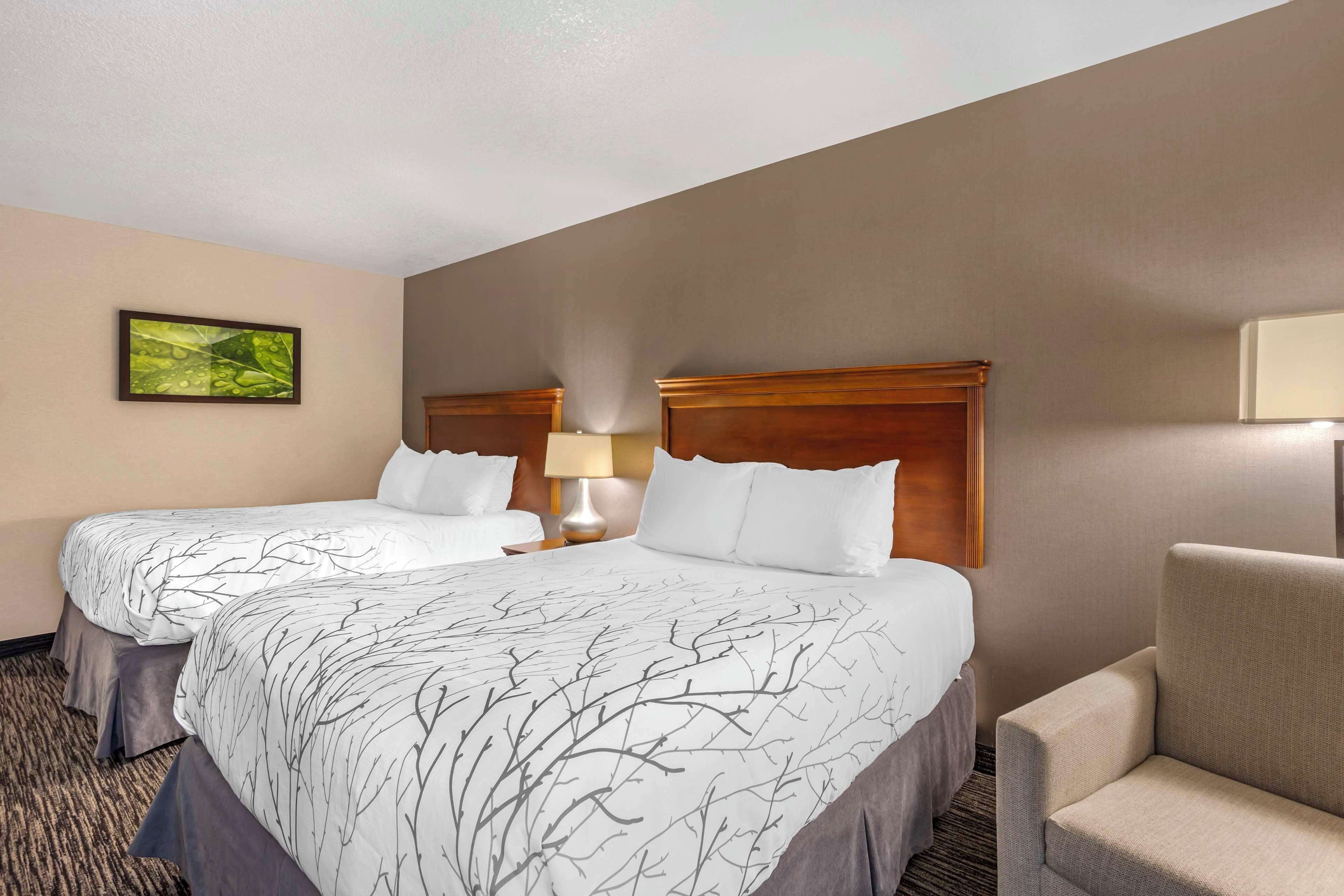 A hotel room with two beds, a chair, a lamp, and a painting on the wall.