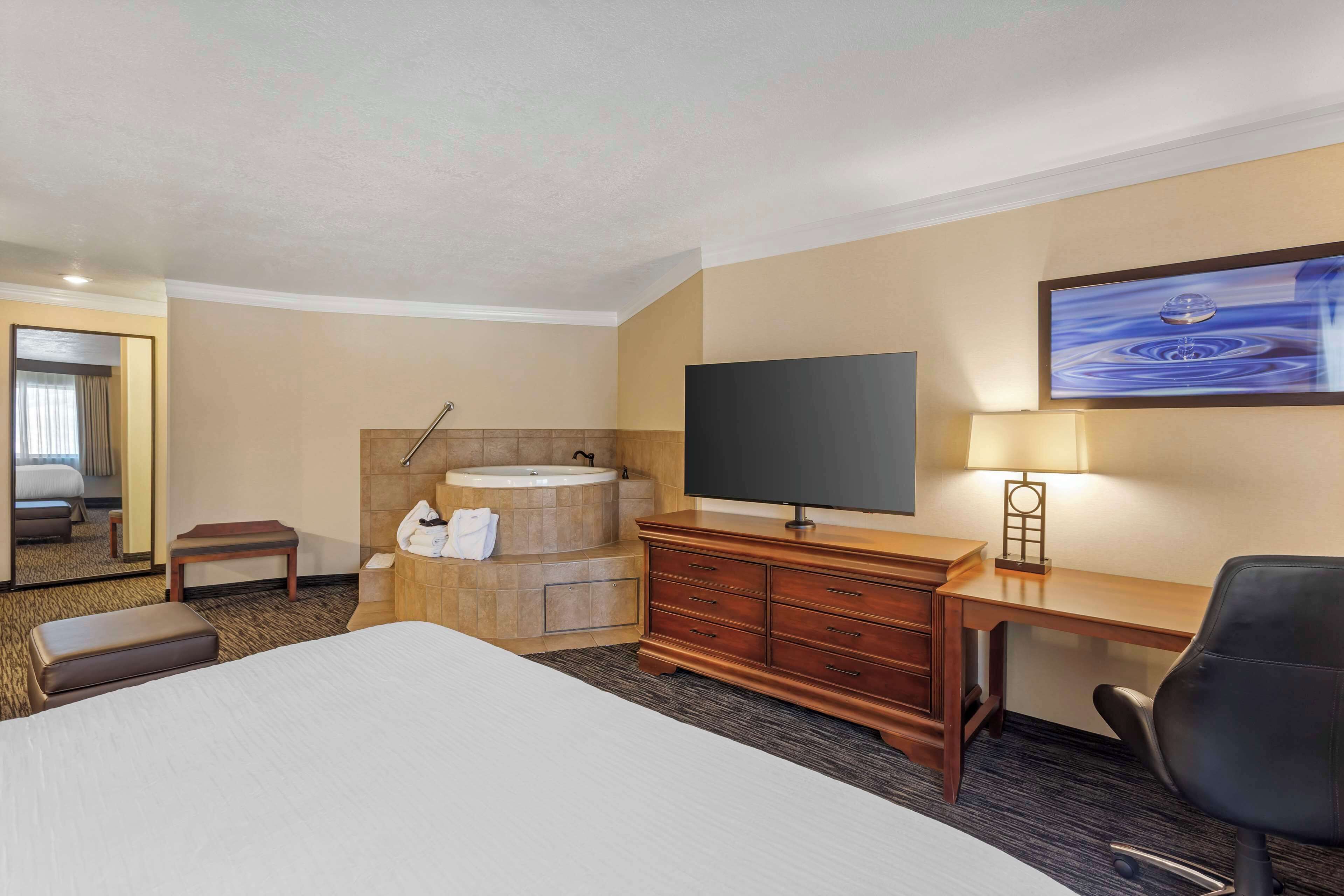 Une chambre d’hôtel avec un grand lit, un bureau en bois, une télévision, une lampe de bureau, une chaise, une salle de bain avec une baignoire et un lavabo, et un tableau accroché au mur.