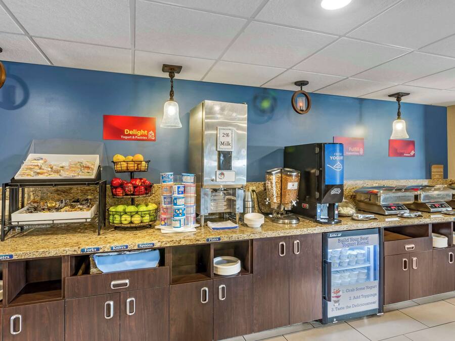 Una estación de desayuno autoservicio con diversos alimentos, un dispensador de leche y un refrigerador para yogur.