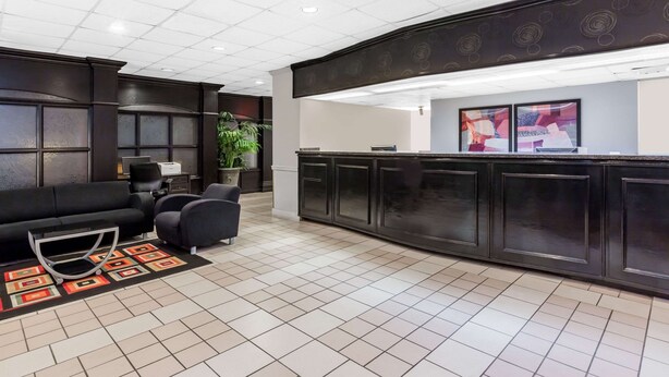 A reception area with a dark reception desk, a black sofa, two armchairs, a potted plant, and a tiled floor. - Days Inn & Suites by Wyndham Jennings (Jennings)