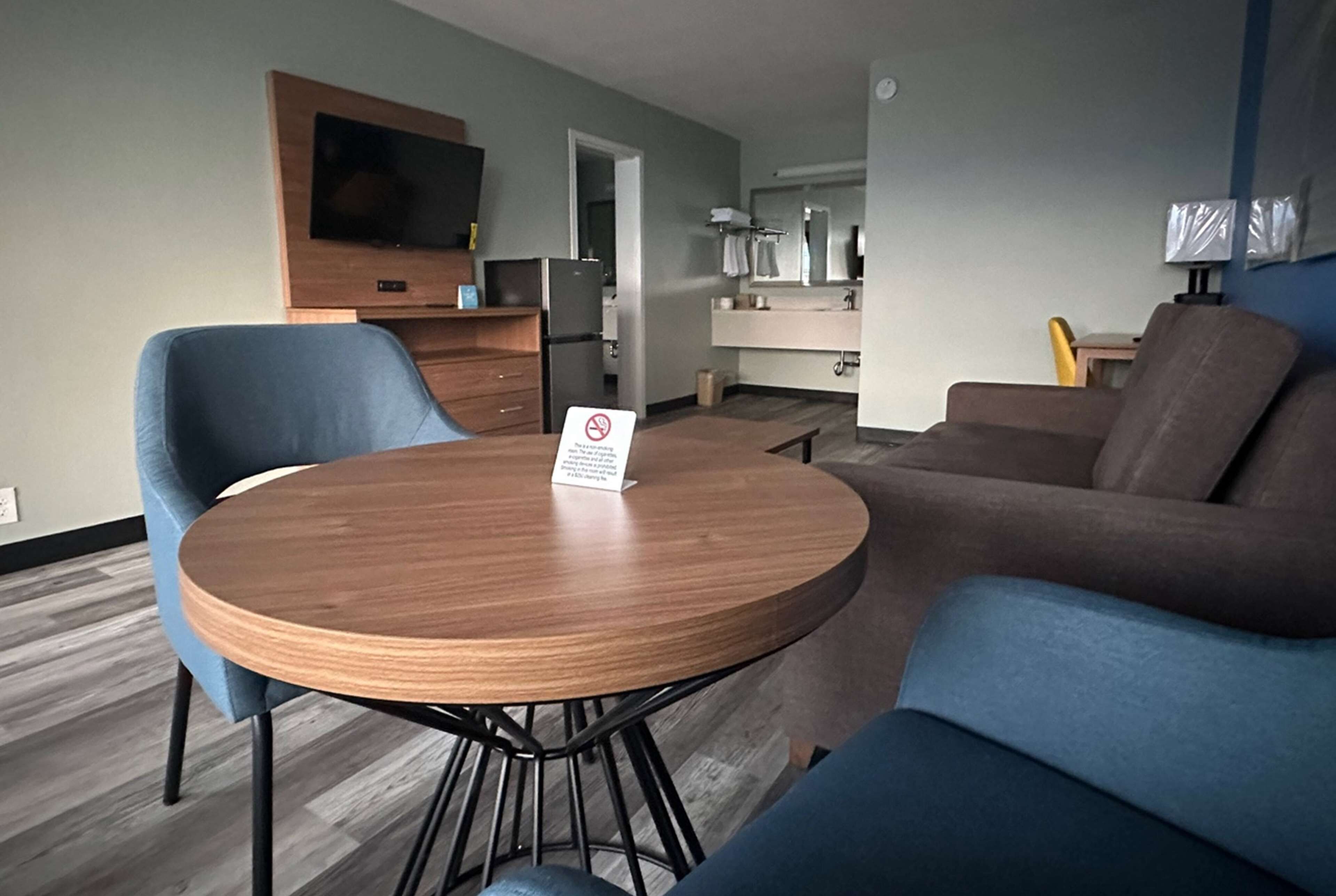 A modern living room with a round wooden table, blue armchairs, a brown sofa, a flat-screen TV mounted on the wall, and a kitchen area in the background.