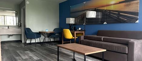A modern hotel room with a wooden floor, a grey sofa, a wooden coffee table, a blue wall with a sunset painting, and a desk with a lamp.