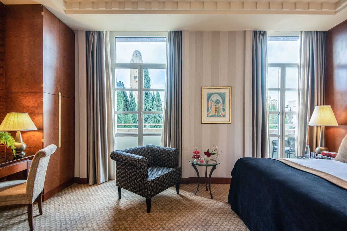 A hotel room with a bed, a chair, a desk, and a small table with a lamp and flowers.