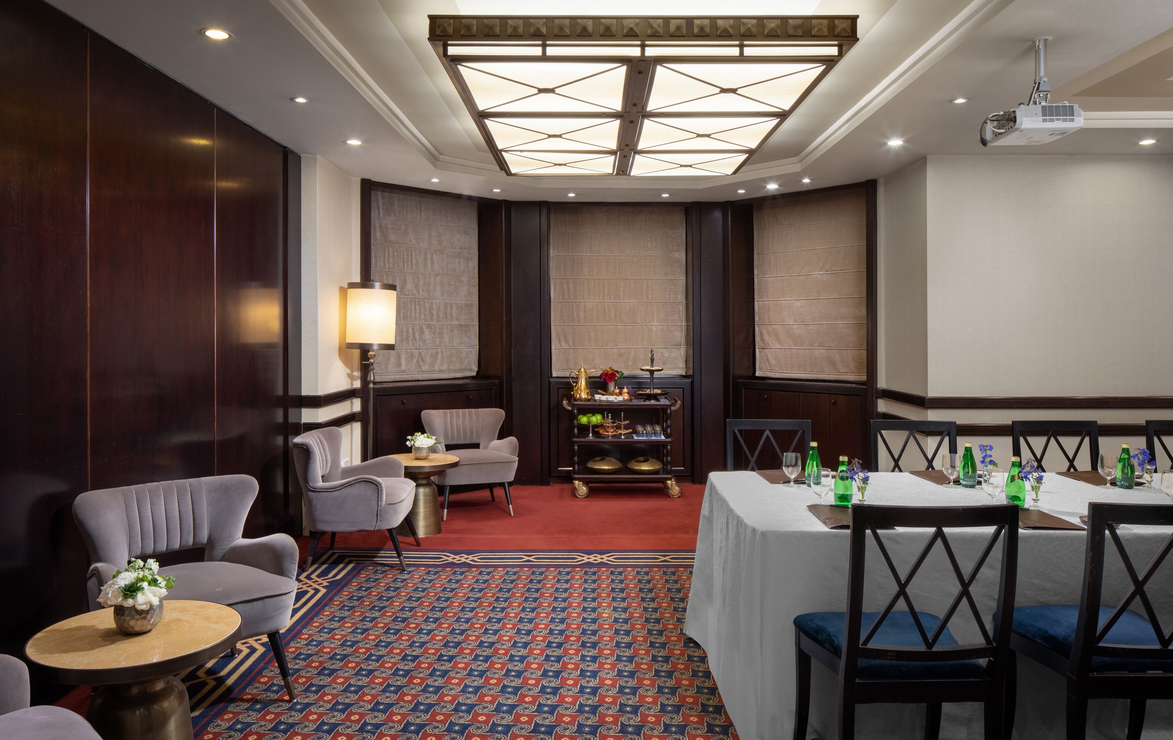 Habitación de hotel con un ventanal grande, una araña, una mesa redonda con sillas, un sofá y un carrito decorativo con objetos.