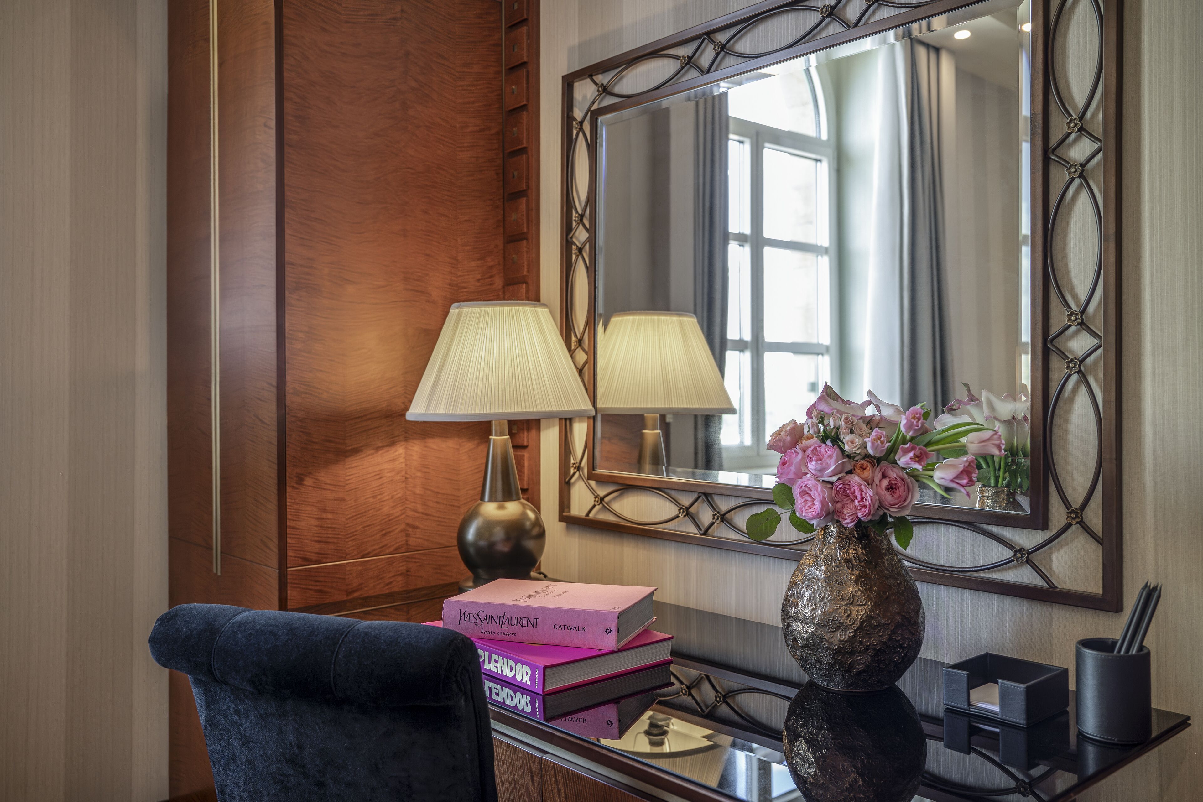 Una habitación de hotel con un panel de pared de madera, un espejo grande, un escritorio con una lámpara, un jarrón con flores y una silla.