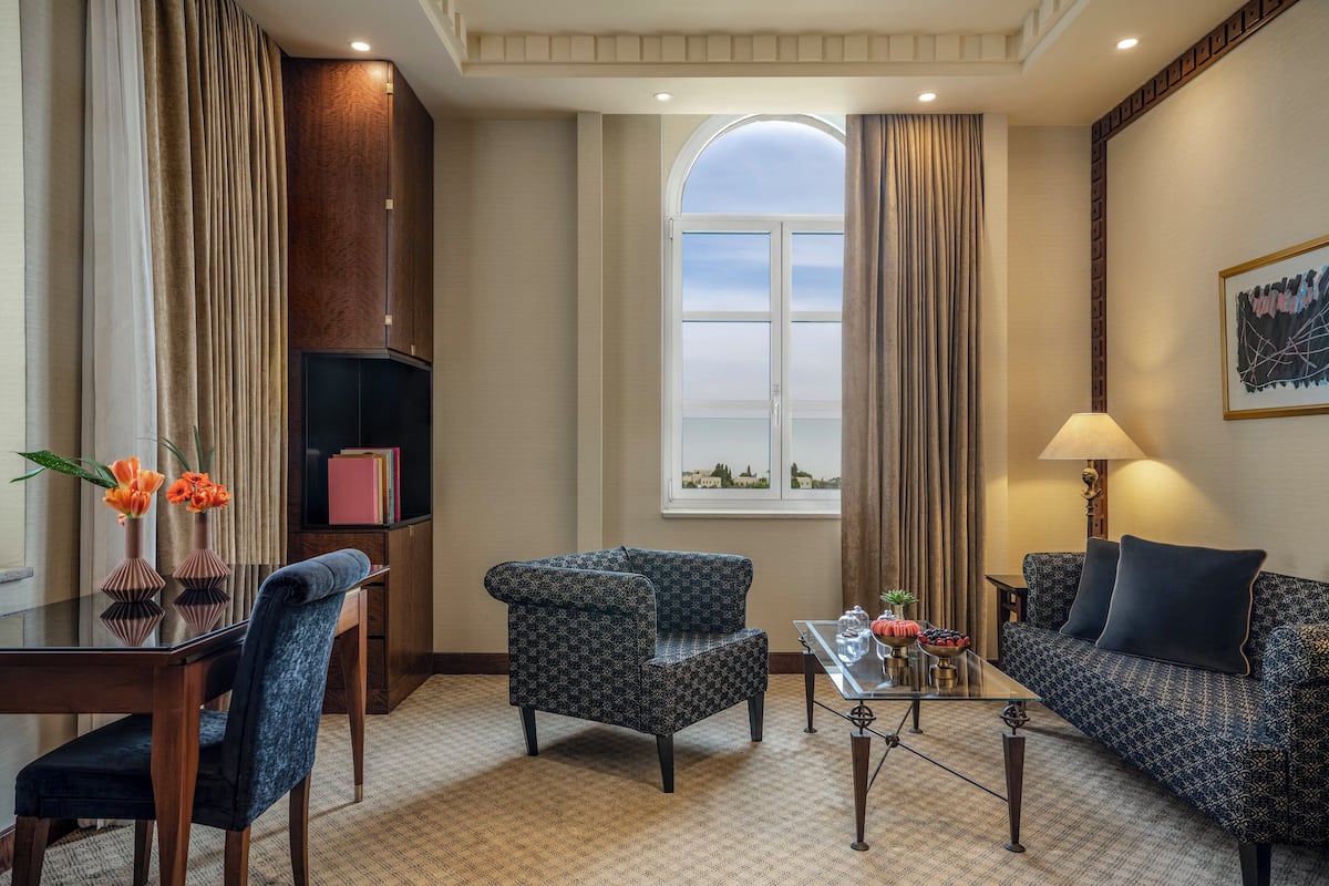 A hotel room with a dining table, two armchairs, a sofa, a small coffee table, and a large window with curtains.
