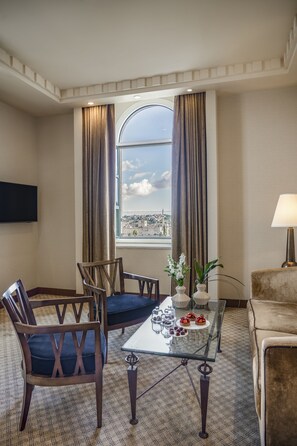 A hotel room with a glass table, two chairs, a sofa, a TV mounted on the wall, and a view of a cityscape through a large window.