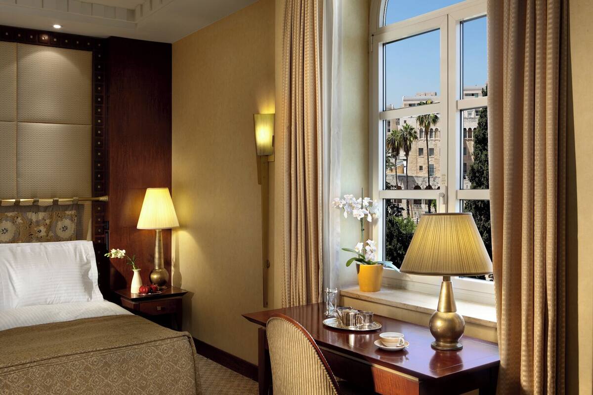 A hotel room with a bed, a desk with a lamp, a chair, a window with curtains, and a view of buildings outside.