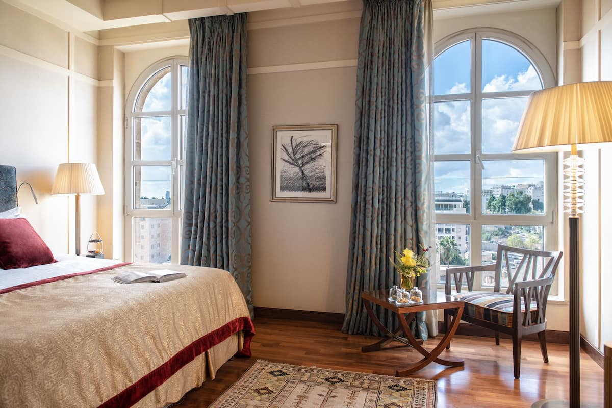 A hotel room with a large bed, a desk with a chair, a window with curtains, and a city view.