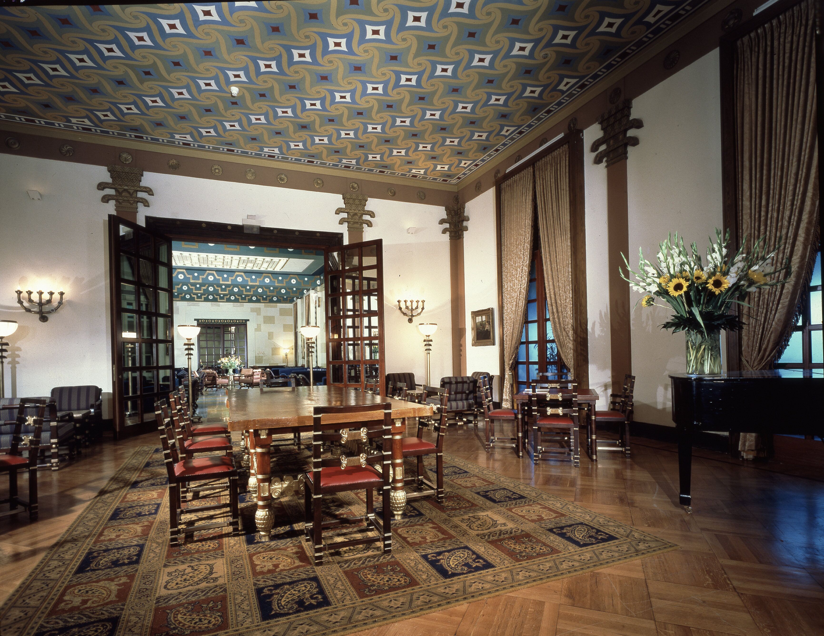 Une vaste pièce avec une grande table et des chaises en bois, un plafond richement décoré et un vase rempli de fleurs.
